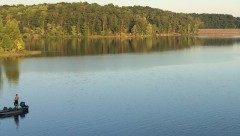 A wide-angle view of a lake with a forested bank in the background. Two men are fishing from a boat on the lefthand side.