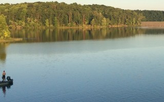 A wide-angle view of a lake with a forested bank in the background. Two men are fishing from a boat on the lefthand side.