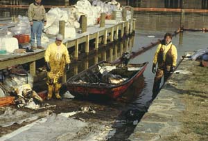 Two mean stand on either side of a boat on a boatlaunch. The men and boat are covered with black diesel fuel. The dock behind the boat has piles of trash in white trash bags.