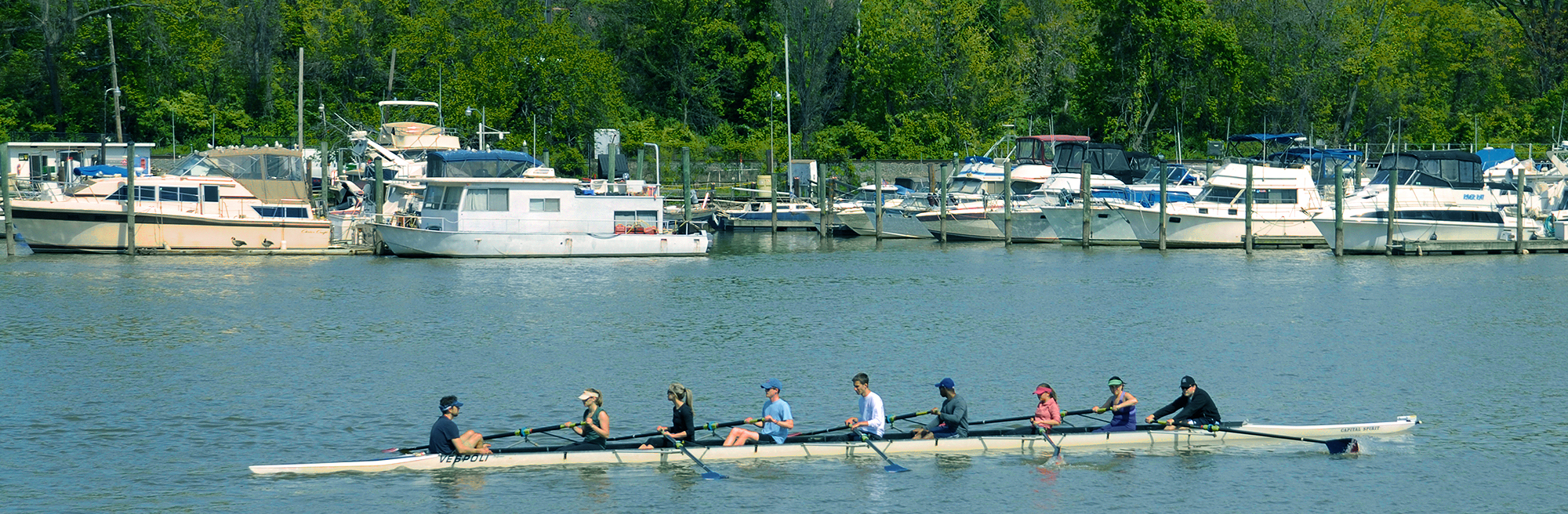 An image of the river with a dock and boats in the background. A long, rowing boat filled rowers in the foreground.