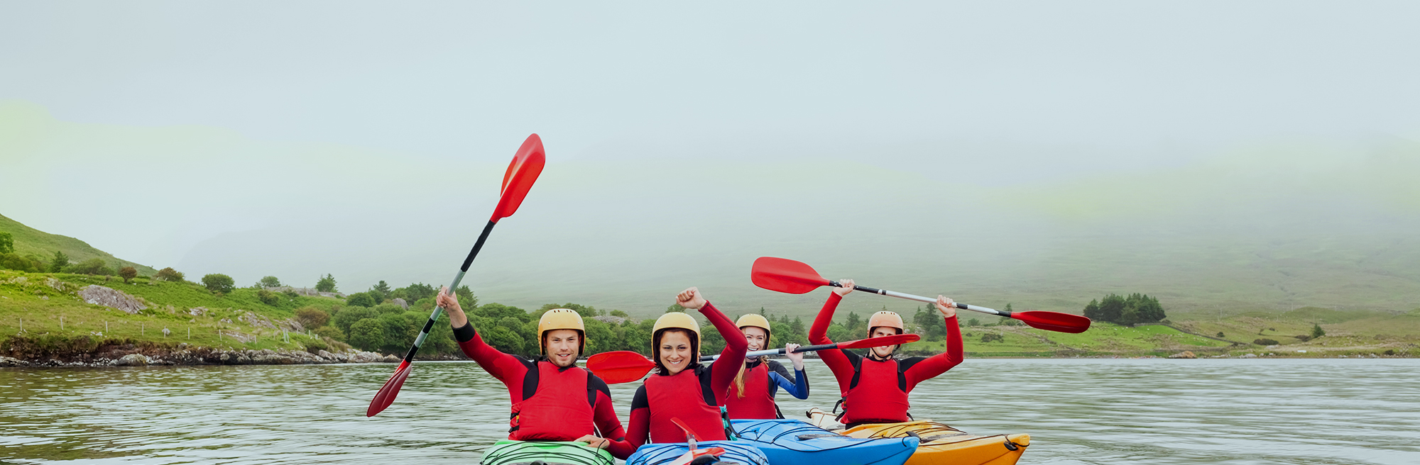 Four people in separate kayaks facing the camera. They have their oars in the air as if celebrating.