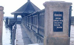 A pier over the river with a sign at the end of the pier that says, "Warning: Avoid contact with polluted river water".