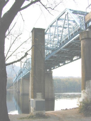 An image of a trestle bridge taken from the shoreline just to the side of the bridge. The view of the of the water and the bottom of the bridge.