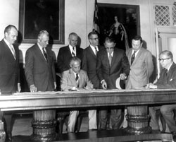 Men stand behind a large table while the ICPRB Compact is signed. 