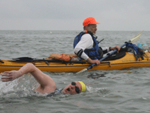 The profile of a kayaker in the background. In front of the kayaker is a simmwer in mid-stroke with his head and right arm out of the water.
