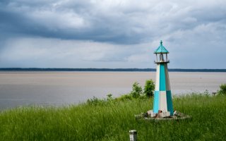 A green, grassy shoreline with a teal and white lighthouse is in the foreground. The river can be seen in the background.