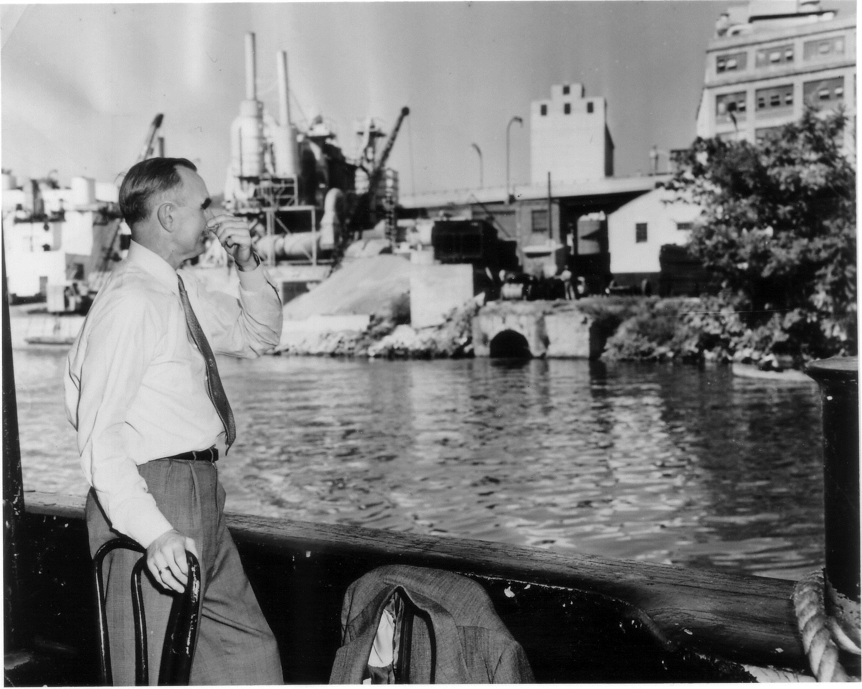 A man stands in a boat, holding his nose. Across the river is an industrial complex with several smokestacks blowing smoke.