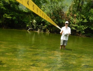 A man standing in the middle of a river. The water is up to his knees. He is holding a measuring tape that stretches to the bank of the river.