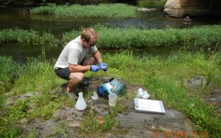 A man crouches next to a stream surrounded by scientific equipment. He has blue plastic gloves on.