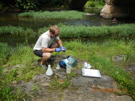 A man crouches next to a stream surrounded by scientific equipment. He has blue plastic gloves on.