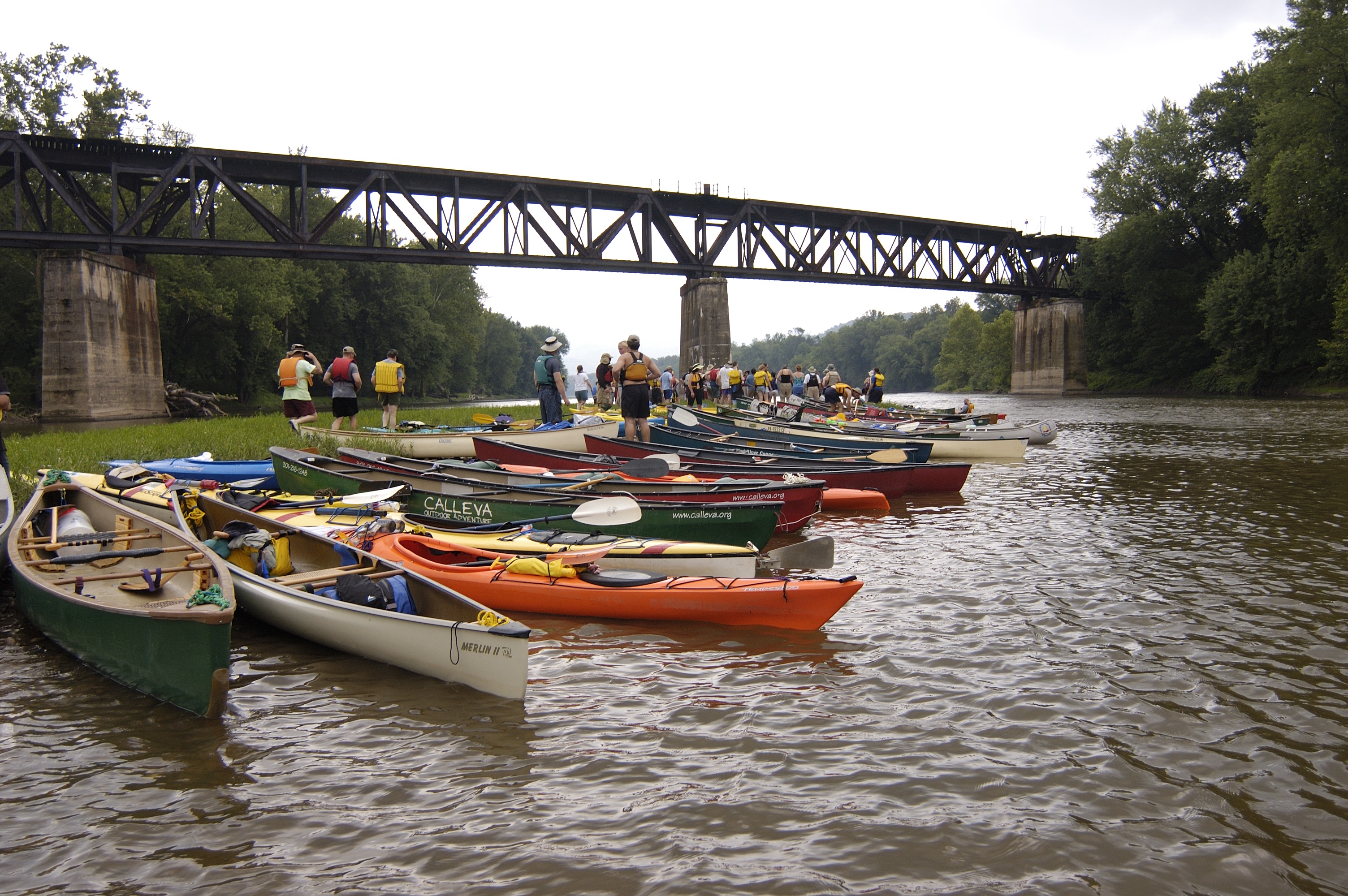 Many kayaks lined up on the shore of a river. A trestle bridge is in the background.
