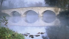 A picture of a bridge across a river. It is slightly foggy. It is a stone bridge with three arches.