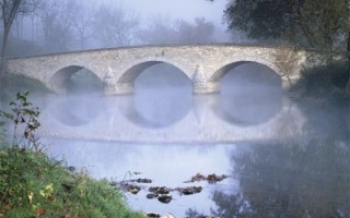 A picture of a bridge across a river. It is slightly foggy. It is a stone bridge with three arches.
