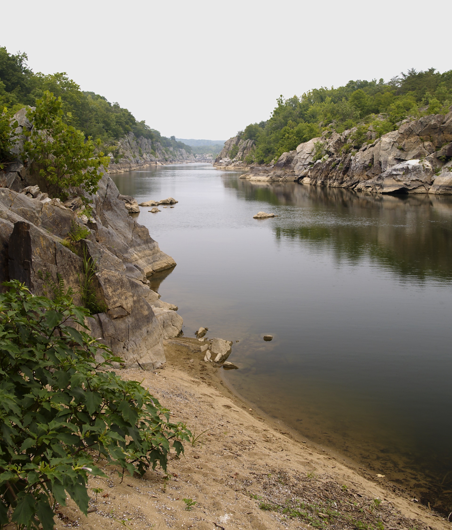 A calm river between two rocky edges. A sandy beach is seen in the foreground.