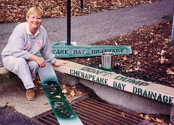 A woman sitting on a curb, posing with a freshly painted storm drain that reads "Don't Dump" and "Chesapeake Bay Drainage".