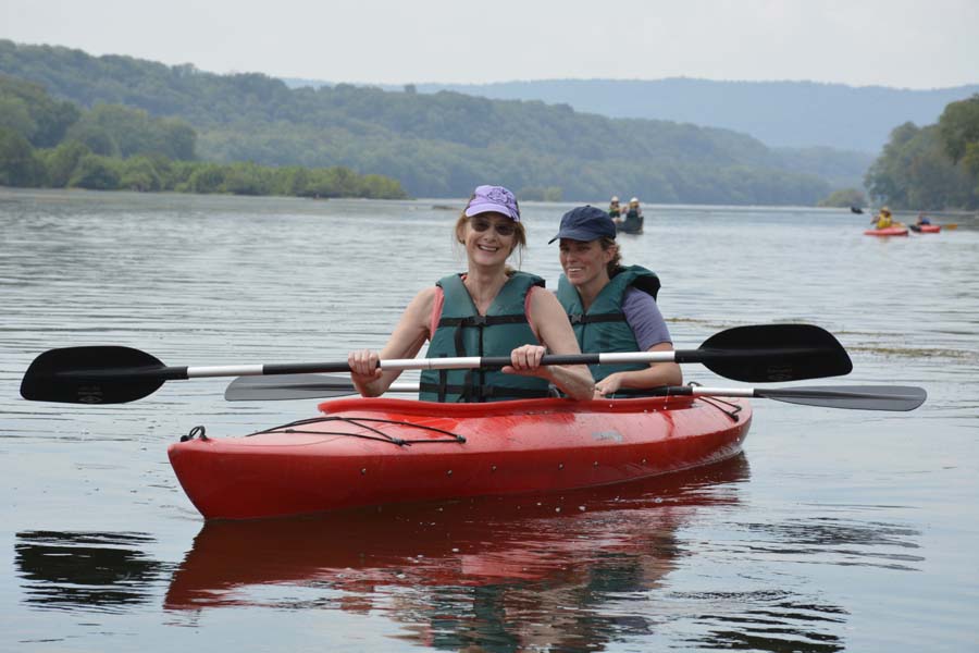 Two women sitting in a red kayak in the middle of the Potomac River. They are each holding a paddle across their lap.