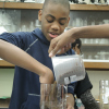A male high school student puts his hand in a beaker while another student pours dirt out of a measuring cup into the beaker.