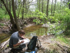 A man kneeling beside a shallow creek. He is writing something down on the clipboard in his hand. A black backpack is in front of him.