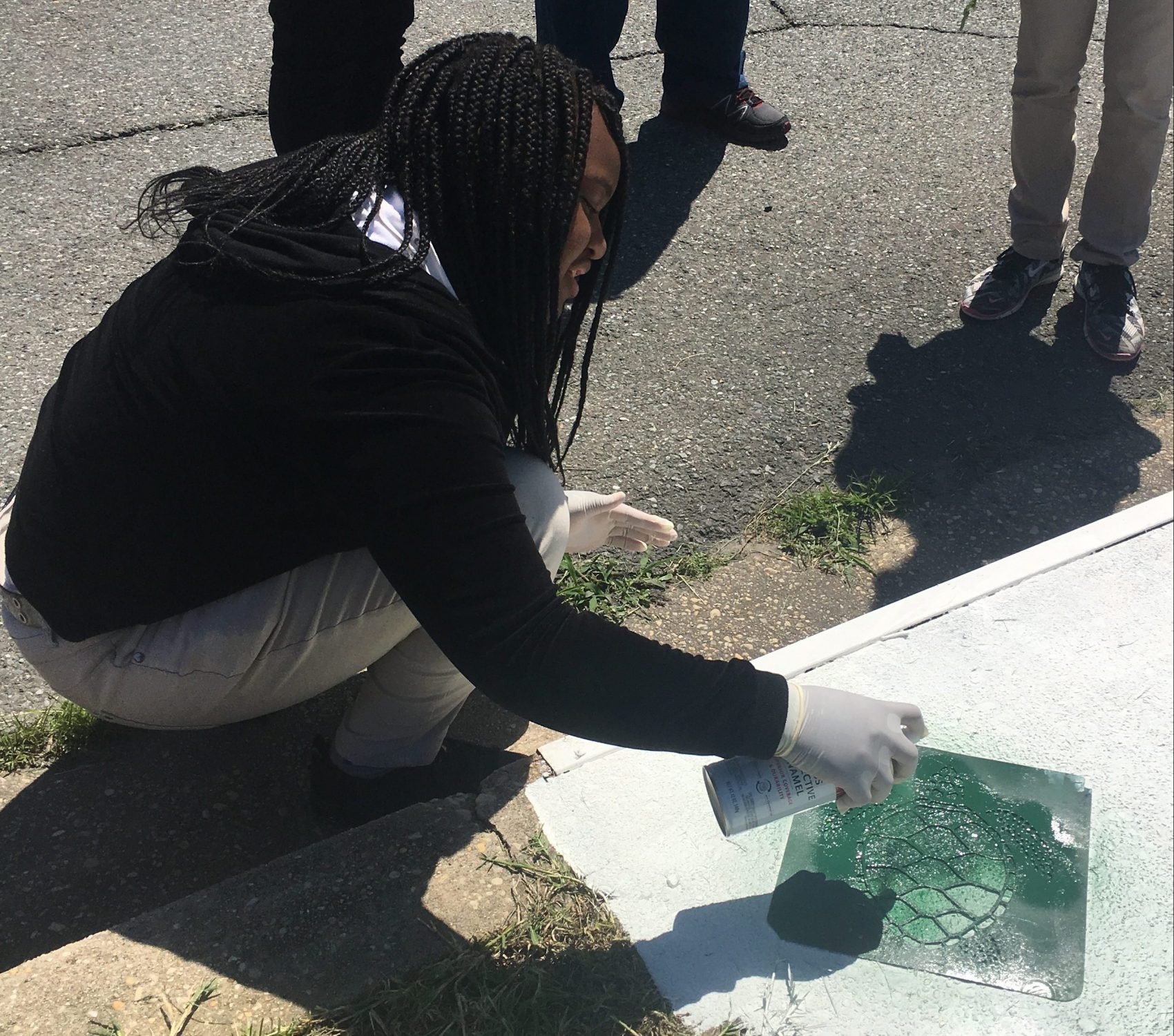 Image of a student kneeling with a can of spray paint in her hand, painting a stormwater drain.