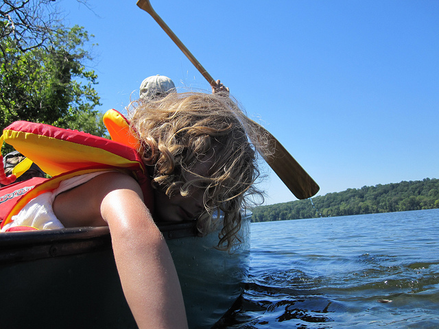 A blond child leaning over a canoe, dragging her hand in the water.