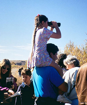 A young girl, sitting on her father's shoulders, with a pair of binoculars.