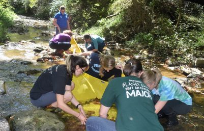 Several people kneeling in a stream, grouped around a net that is collecting benthic macroinvertebrates.