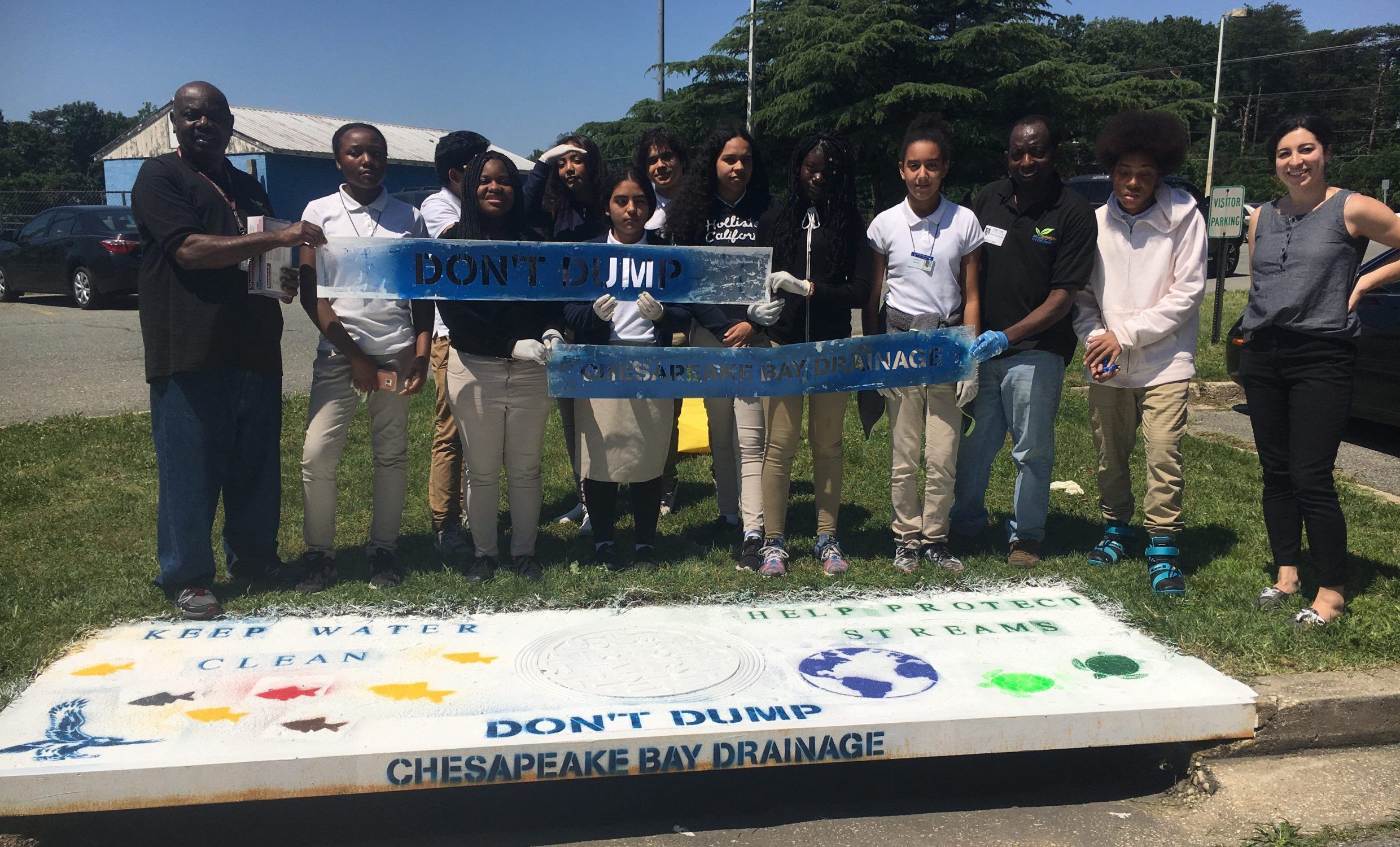 Several students standing behind a freshly painted stormwater drain. They are holding stencils that say "Don't Dump" and "Chesapeake Bay Drainage".