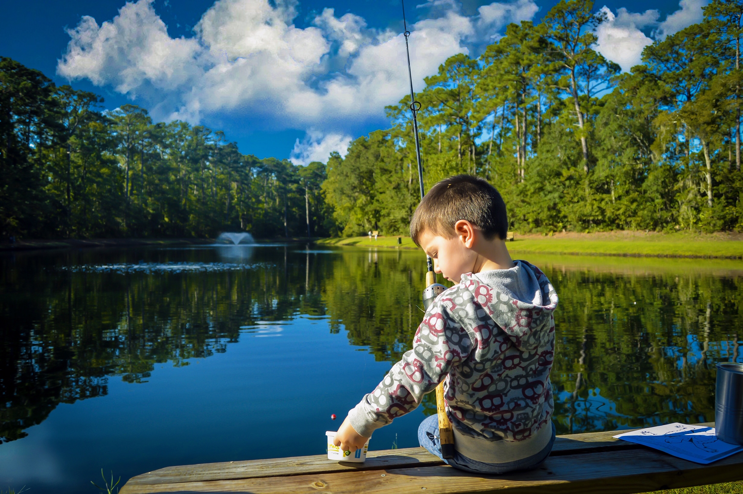 Little boy sitting on a bench reaching for bait. A lake is in the background.