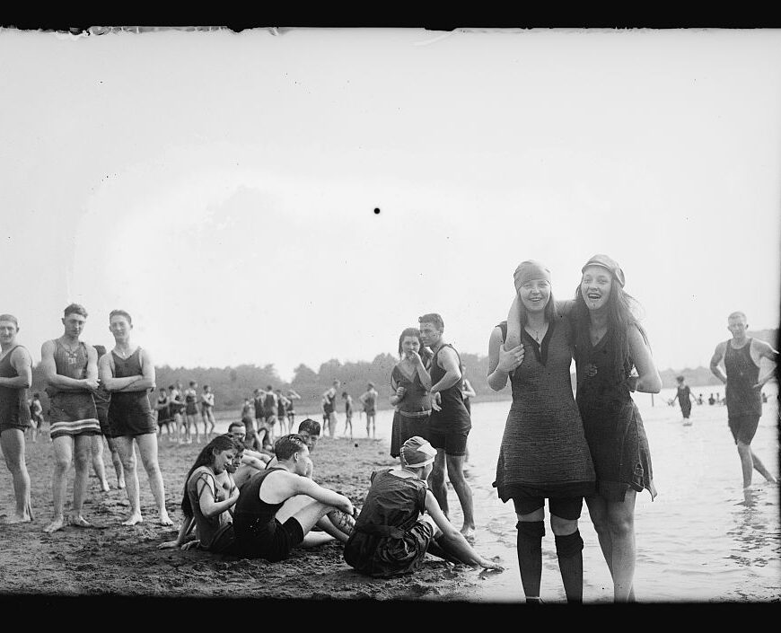 Two women stand in the sand along the tidal basin / Potomac River among other bathers.