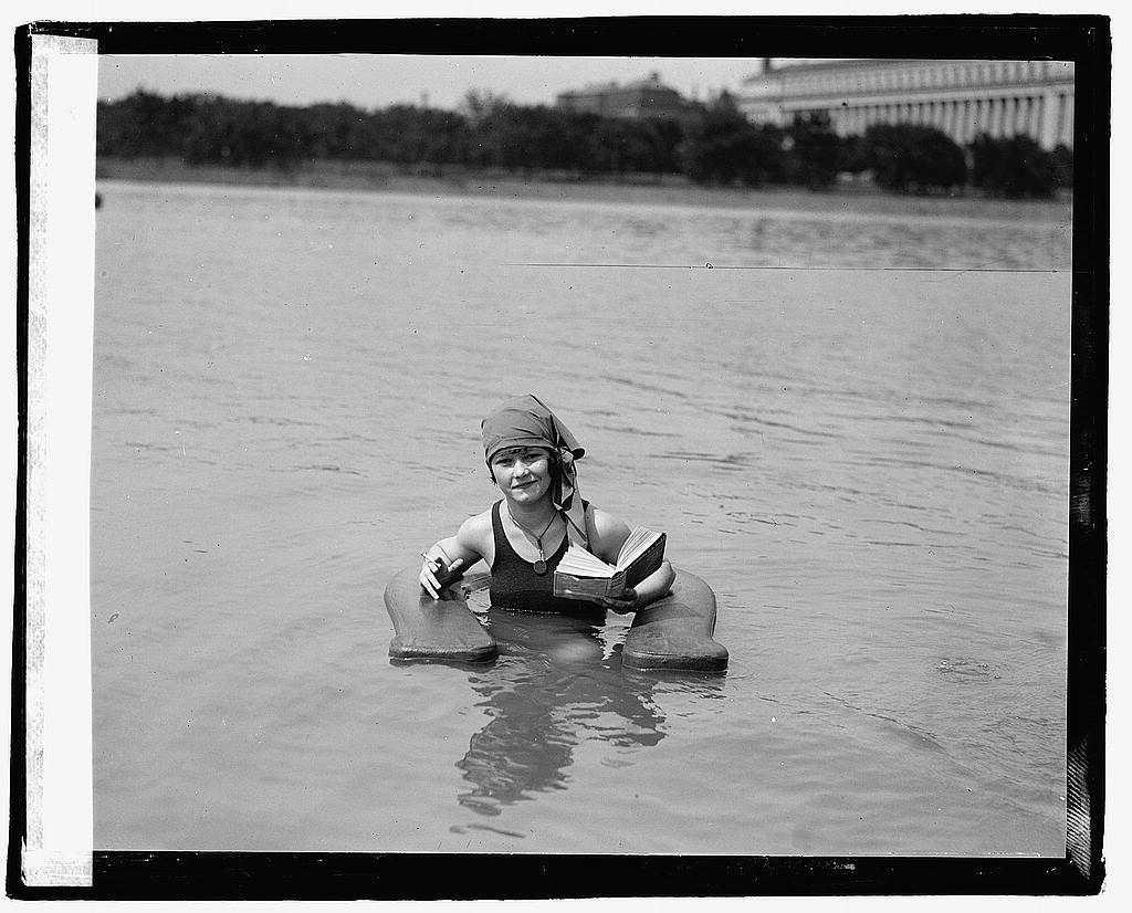 A woman sits and reads in the water in an old fashioned bathing suit.