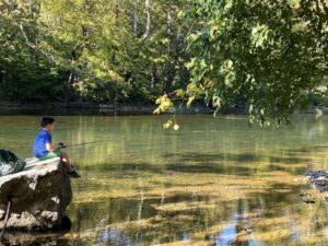 Young boy with a fishing pole sitting on a rock in the Shenandoah River. 