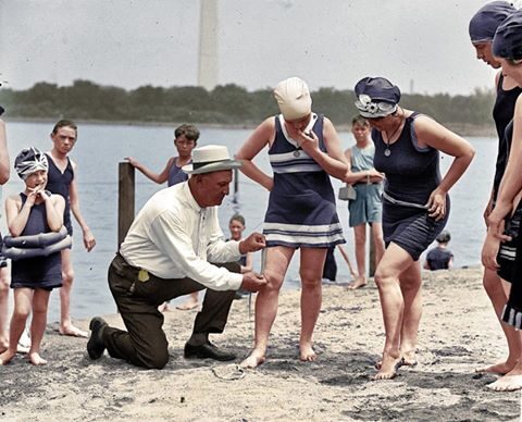 Women in old fashioned bathing suits are having the length of their suits measured while standing at the beach on the Tidal Basin
