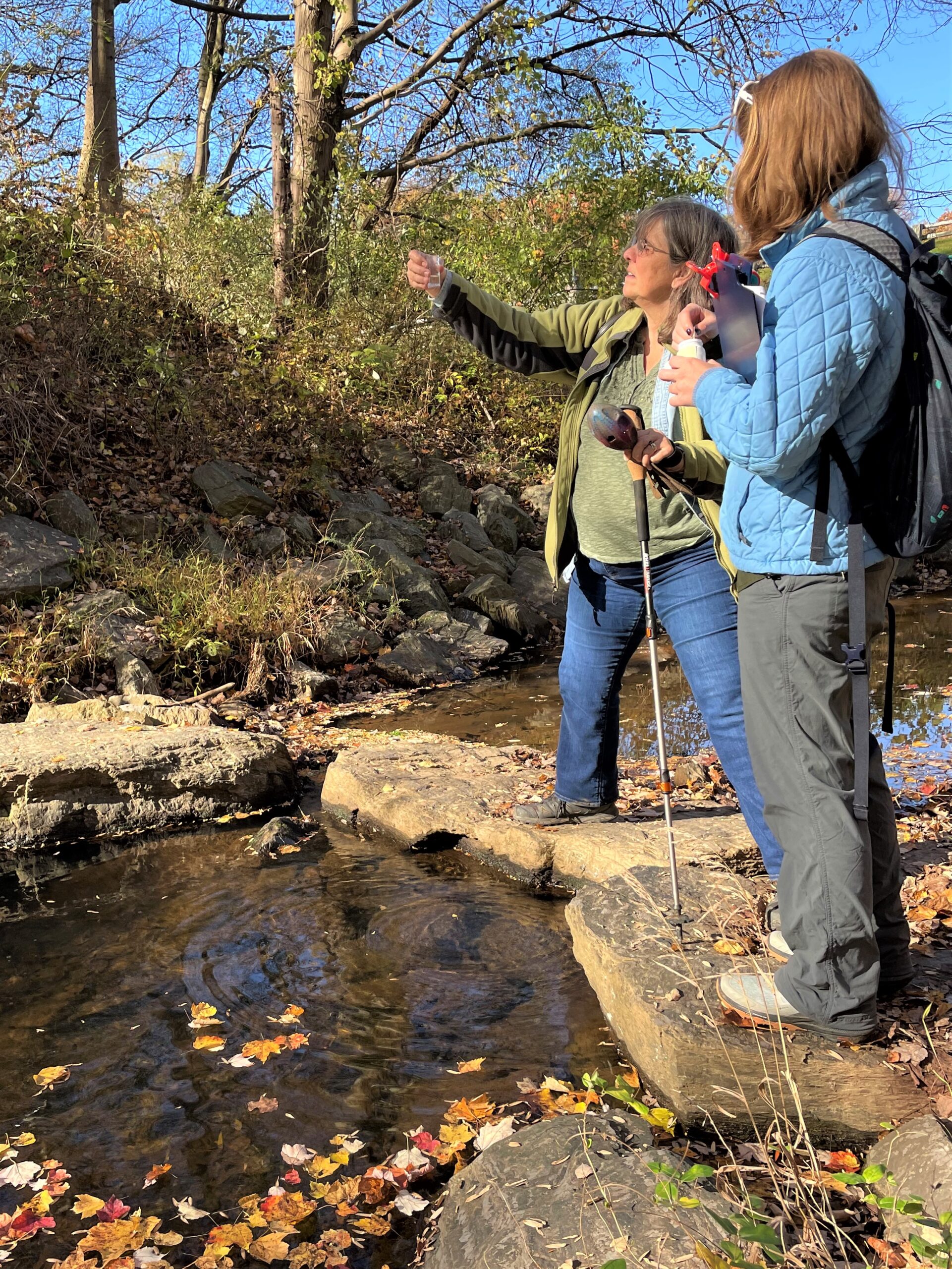 Two people stand alongside a stream. One person is holding a water sample up.